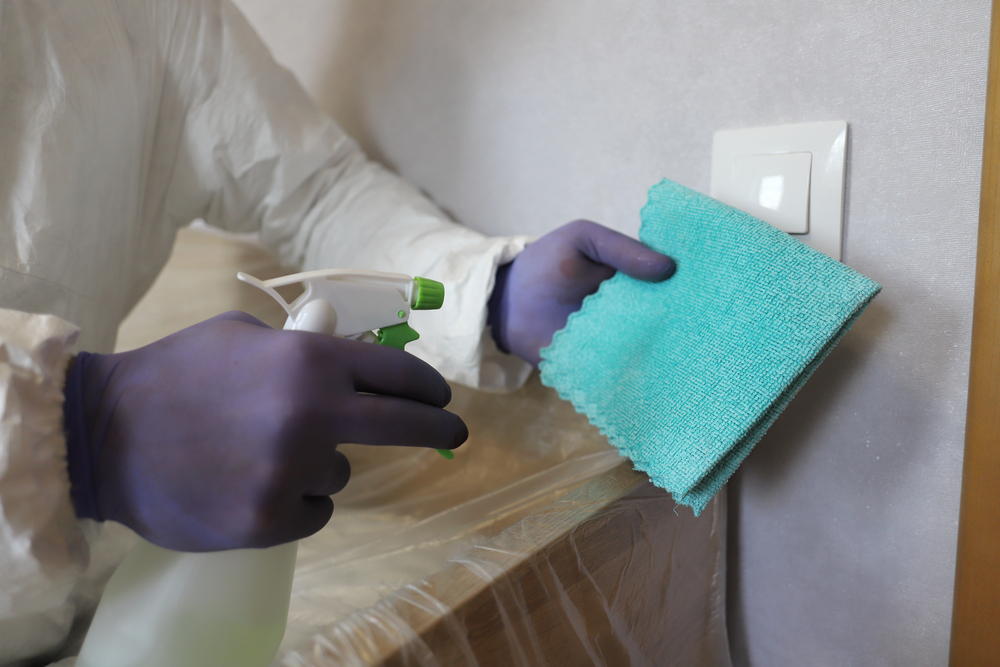 Mold inspector in protective suit and gloves sanitizing a light switch with spray bottle and cloth